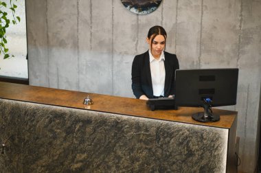 Modern otel lobisinde genç profesyonel resepsiyonist. Resepsiyonları kontrol ediyor, gülümsüyor ve bilgisayar kullanıyor.