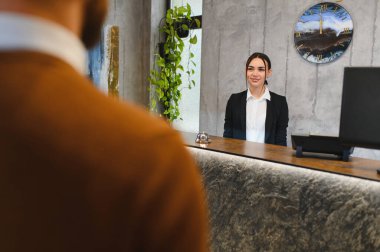 Tezgahta duran gülümseyen genç resepsiyonist, bulanık bir müşteriyle iletişim kuruyor, otel resepsiyonunda hizmet veriyor.