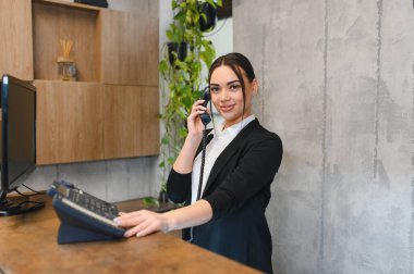 Kendine güveni tam bir kadın resepsiyonist. Tezgahta duruyor. Ofis telefonunda konuşurken kameraya gülümsüyor.