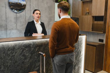 Kadın otel resepsiyonisti gülümsüyor, modern lobi resepsiyonundaki odasına giriş yapan bir erkeğe yardım ediyor.
