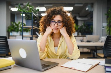 Bayan öğrenci dizüstü bilgisayarla çalışırken yorgun ve sıkılmış hissediyor, modern bir ofiste eğitim görevleriyle boğuşuyor.