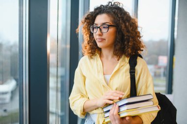 Kıvırcık saçlı, gözlük takan, kitap taşıyan ve sırt çantası olan genç bir kız öğrenci. Üniversite koridorunda pencerenin kenarında durup ders çalışıyor.