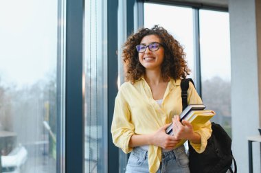 Kıvırcık saçlı ve gözlüklü genç bir kız öğrenci, modern bir binada ders kitabı ve sırt çantası tutarken gülümsüyor.