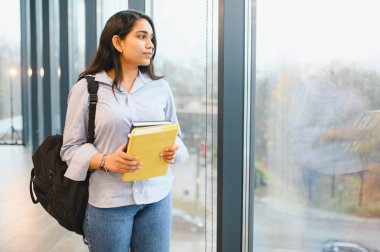 Sırt çantası takan ve kitap tutan Hintli bir öğrenci, büyük bir pencerenin yanında duruyor ve düşünceli bir şekilde dışarı bakıyor.