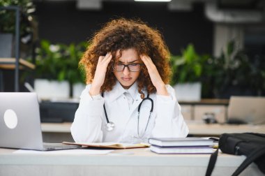 Laboratuvar önlüğü ve steteskop giyen genç bayan tıp öğrencisi yoğun online çalışmalar sırasında baş ağrısı ve stresten muzdarip.