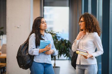 Üniversite kampüsünde iki farklı kız öğrenci kitap ve sırt çantalarıyla sohbet ediyorlar.