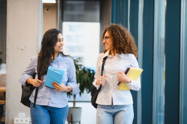 Yürüyen ve konuşan iki gülümseyen kız öğrenci, kitap ve sırt çantaları taşıyorlar, arkadaşlığı ve modern eğitimi temsil ediyorlar.