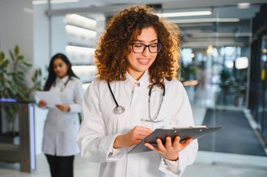 Steteskop ve gözlük takan genç bir kadın doktor, modern bir hastane ortamında gülümseyen ve danışmanlık yapan hasta sağlığı bilgileri