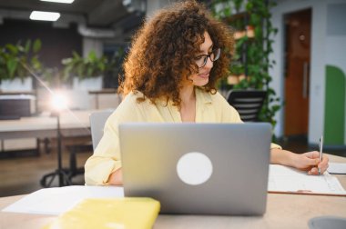 Kıvırcık saçlı ve gözlüklü genç bir kadın modern bir ofiste çalışıyor, notlar yazıyor ve dizüstü bilgisayar kullanıyor.
