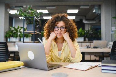 Genç kız öğrenci çevrimiçi eğitime odaklanmış, kitaplarla dolu modern bir çalışma ofisinde dizüstü bilgisayar eğitimi görüyor.