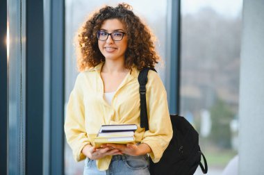 Genç bir kız öğrenci ayakta, elinde kitaplarla modern bir üniversitede ya da üniversite ortamında sırt çantası taşıyor.