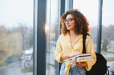 Camın kenarında duran kıvırcık saçlı ve gözlüklü genç bir kadın. Ders sırasında kitap ve sırt çantası tutuyor.
