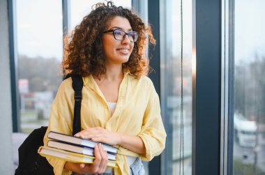 Gözlük ve sırt çantası takan, ders kitaplarını tutan, geniş pencerenin yanında duran genç bir kız öğrenci. Öğrenme, eğitim ve gelecekteki başarı kavramı