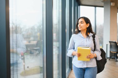 Sırt çantası ve kitapları olan Hintli bir öğrenci modern bir üniversite kütüphanesinde yürüyor, başka tarafa bakıyor, eğitime hazır.