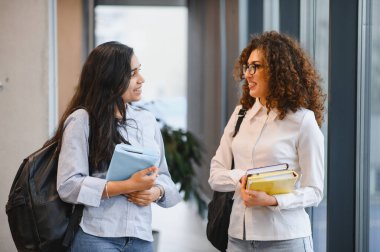 Modern bir üniversite koridorunda birlikte yürüyen, kitap ve sırt çantaları taşıyan, konuşan ve gülümseyen iki farklı genç kız öğrenci.