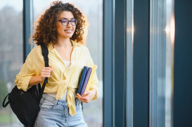 Genç kıvırcık saçlı, gözlüklü ve sırt çantalı bir öğrenci elinde renkli kitaplarla üniversite koridorunda dikilip gözlerini kaçırıyor.