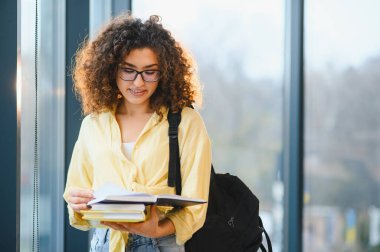 Ders çalışan, gözlüklü, sırt çantalı, elinde kitap tutan bir kız öğrenci. Eğitim, öğrenme ve akademik hedefleri temsil eder