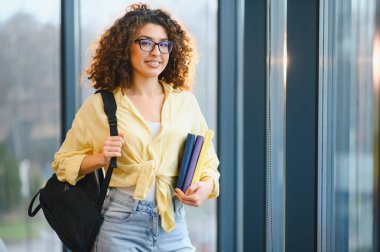 Modern bir binada kameraya gülümserken elinde kitaplarla sınıfa giden mutlu bir kız öğrenci.