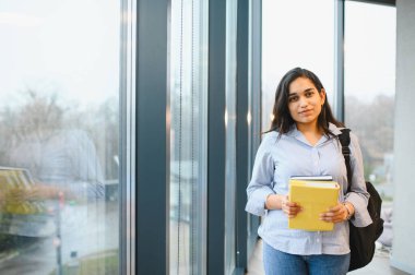 Genç Hintli kız öğrenci elinde kitaplarla kampüs binasının içinde büyük bir koridorda duruyor.