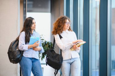 Sırt çantaları ve kitaplar taşıyan iki farklı kız öğrenci modern bir üniversite binasında duruyor ve ders için hazırlanıyorlar.