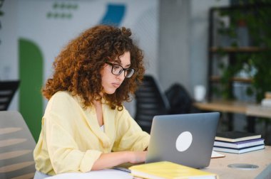 Kıvırcık saçlı ve gözlüklü genç bir kadın eğitim ya da iş için dizüstü bilgisayar kullanıyor. Uzaktan öğrenme ve dijital okur yazarlığa odaklan