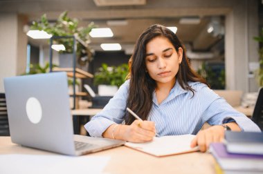 Genç Hintli kadın, modern ofis ortamında dizüstü bilgisayar üzerinde çalışırken, bir deftere notlar yazmaya odaklandı.