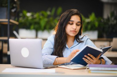 Hintli öğrenci öğrenme ve derslerine konsantre olma, çevrimiçi eğitim için dizüstü bilgisayar kullanırken kitap okuma