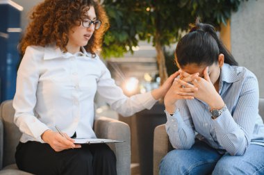 Kadın psikolog, terapi seansı sırasında yüzünü kapatan stresli Hintli bir kadına empati ve destek sağlıyor.