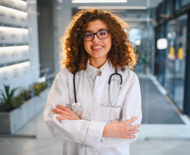 Kıvırcık saçlı, gözlüklü, laboratuvar önlüğü ve steteskop takan, modern bir klinikte güvenle gülümseyen genç bir bayan doktor.