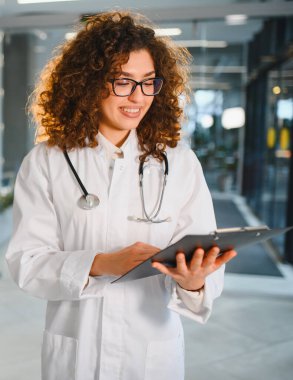 Kıvırcık saçlı, gözlüklü, laboratuvar önlüğü ve steteskop takan genç bir kadın doktor, panoya yazarken gülümsüyor.