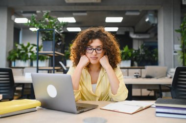 Kıvırcık saçlı genç bir kadın dizüstü bilgisayarla çalışırken yorgun düşüyor, uzun saatler sonra tükeniyor.