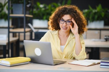 Kıvırcık saçlı ve gözlüklü genç bir kadın çevrimiçi kursa odaklanıyor, modern bir ofiste öğreniyor veya uzaktan çalışıyor.