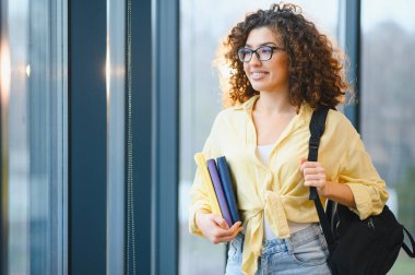 Sırt çantası ve kitaplarla güvenle gülümseyen genç bayan öğrenci, parlak bir akademik geleceğe ve yeni bilgilere bakıyor.