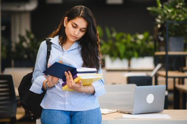 Kitaplar taşıyan genç Hintli bir kadın, modern eğitim ortamında öğrenme ve öğrenim görürken dizüstü bilgisayarla odaklanmış görünüyor.