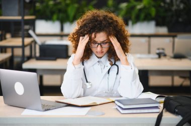 Laboratuvar önlüğü ve steteskop giymiş bir tıp öğrencisi. Çalışırken başını stresten ve yorgunluktan tutuyor.