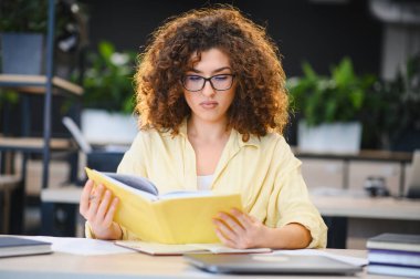 Genç kız öğrenci, modern bir kütüphane veya ofis alanında ders çalışmaya, ders kitabı okumaya ve öğrenme materyallerine odaklandı.