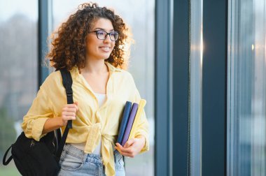 Sırt çantası ve kitap taşıyan kıvırcık saçlı gülümseyen genç bir kadın, modern bir binada duruyor, öğrenmeye ve eğitime hazır.