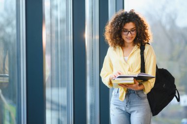 Sırt çantası ve ders kitaplarıyla çeşitli genç bayan öğrenciler parlak, modern bir üniversite koridorunda okuyor, gülümsüyor ve öğrenmeye odaklanıyor.