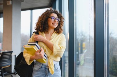 Gülümseyen, pencereden dışarı bakan genç bir kadın. Elinde kitaplar var ve bir sırt çantası takıyor. Eğitim ve üniversite hayatını temsil ediyor.