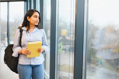 Not defterleri ve sırt çantası taşıyan Hintli genç bir kız öğrenci modern bir üniversite binasının koridorunda yürüyor.