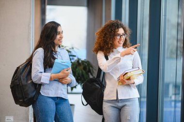 Sırt çantası ve kitap taşıyan, konuşan ve işaret eden iki gülümseyen kız öğrenci modern bir üniversite binasında yürürken. Eğitim ve kampüs hayatının bir sembolü.