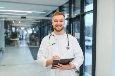 Laboratuvar önlüğü ve steteskop giymiş gülümseyen erkek doktor, modern bir klinikte veya hastanede elinde bir not panosu ve kalem tutuyor.