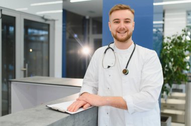 Genç bir erkek doktor resepsiyonda duruyor, kendine güvenerek gülümsüyor ve kameraya bakıyor, üzerinde tıbbi bir ceket ve steteskop var.