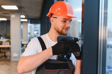 Modern bir ofiste kablosuz matkapla pencere iskeleti inşaatında veya muhafazasında çalışan genç bir inşaatçı.