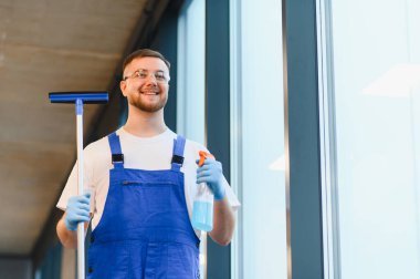 Mavi üniformalı, koruyucu gözlüklü ve eldivenli gülümseyen adam büyük pencerelerin yanında temizlik spreyi ve cam sileceği tutuyor.