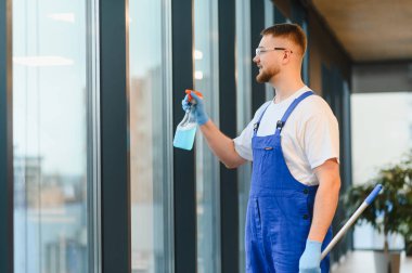 Erkek temizlikçi, tulum giyiyor ve güvenlik gözlüğü takıyor. Geniş ofis penceresine sprey sıkarak bakım sağlıyor.