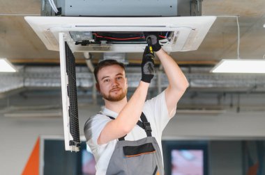 İş eldivenli nitelikli genç adam modern bir ofiste bir klima kasetinin bakımını yapıyor.