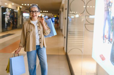 Gülümseyen, alışveriş torbaları tutan, modern bir alışveriş merkezinde yürüyen neşeli bir son sınıf kadını. Tüketim ve yaşam tarzı kavramı