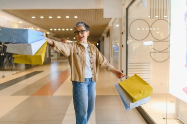 Alışveriş torbalarıyla gezen ve alışveriş merkezinde gülümseyen yetişkin bir kadın satın alma ve tüketim yaşam tarzından zevk alıyor.