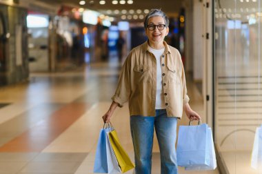 Alışveriş torbaları taşıyan mutlu, modern bir alışveriş merkezinde yürürken gülümseyen, tüketimden zevk alan yaşlı bir kadın.